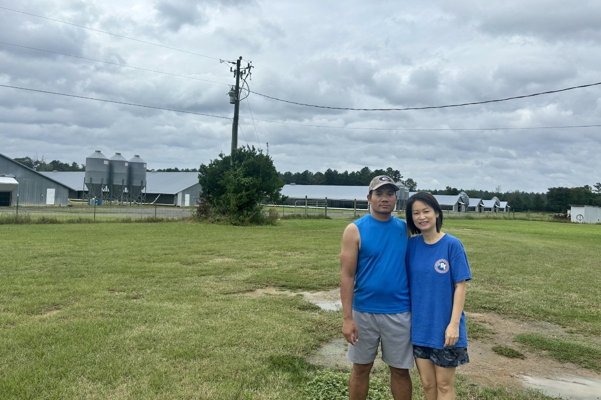 A Vietnamese American couple wearing blue t-shirts stands in a large green field on a chicken farm