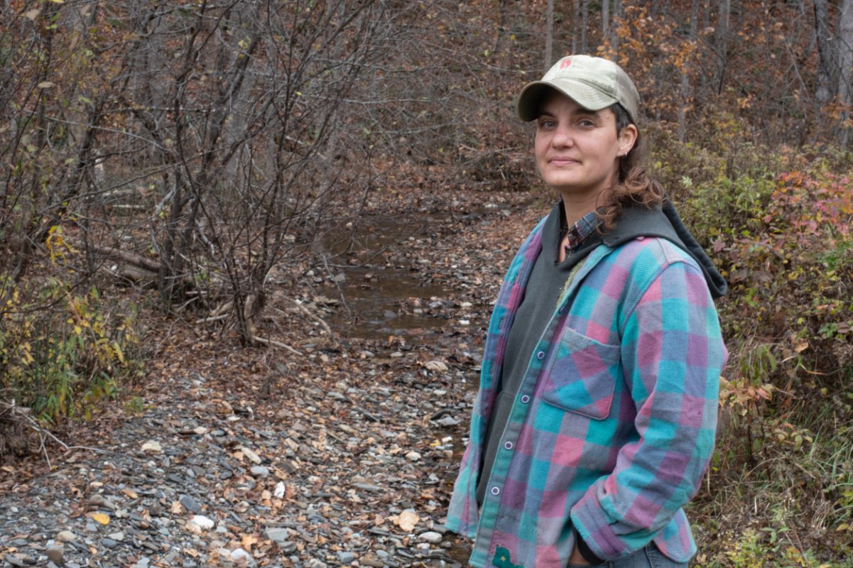A woman wearing flannel and a cap is a farmer, standing in front of the woods