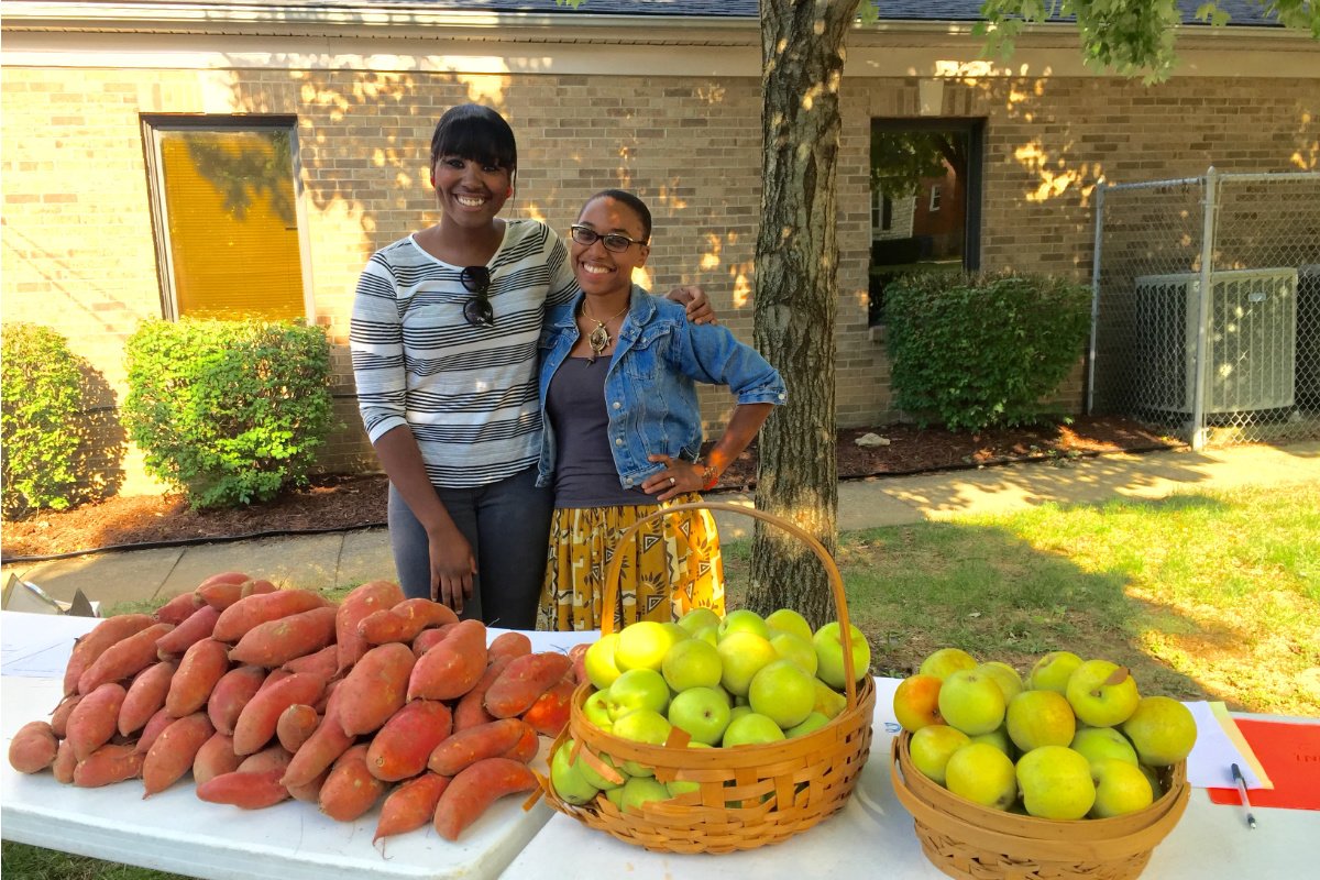 Two Black women stand behind an outdoor tale with fresh sweet potatoes and fruits in baskets