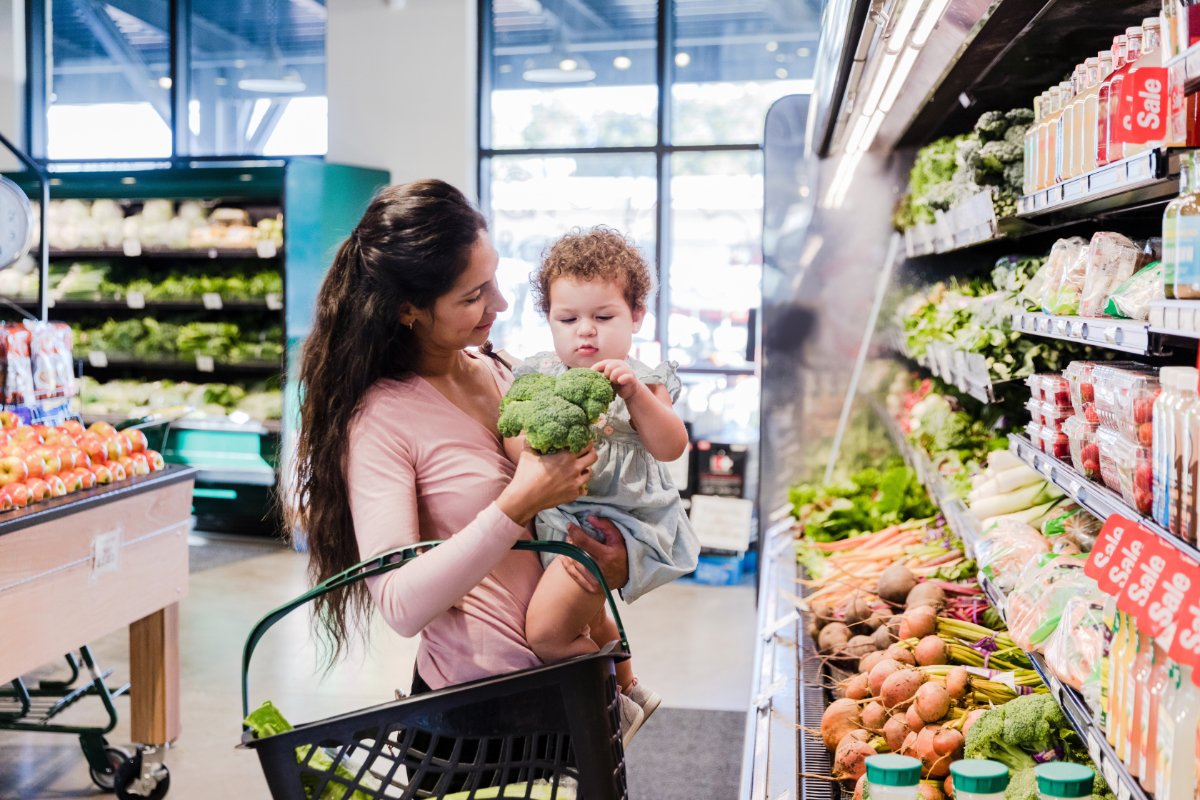 A woman carrying a toddler shows the toddler a broccoli inside a grocery store