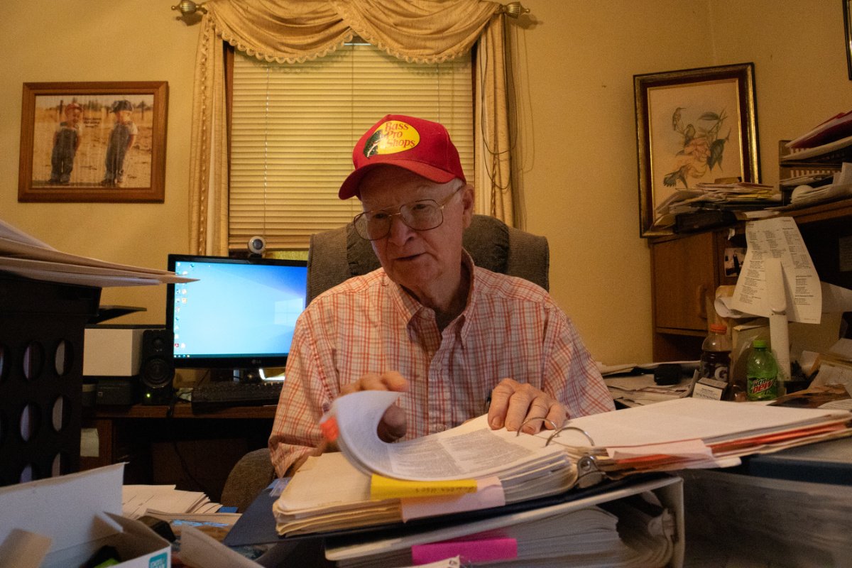 An elder man wearing a bass pro shop red cap sits at a table reading through documents