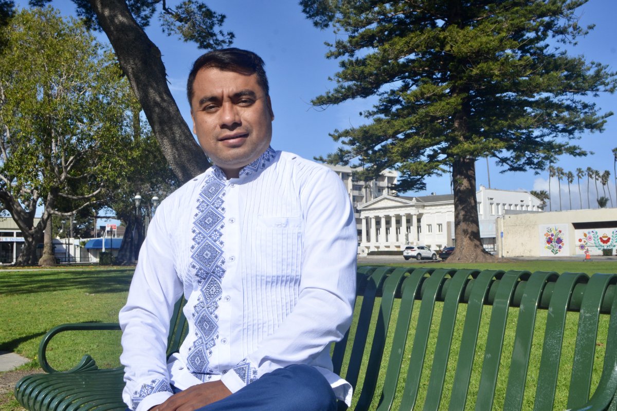 An Indigenous Mixtec man sits on a public green bench in a park, dressed in a white shirt with blue emboidery