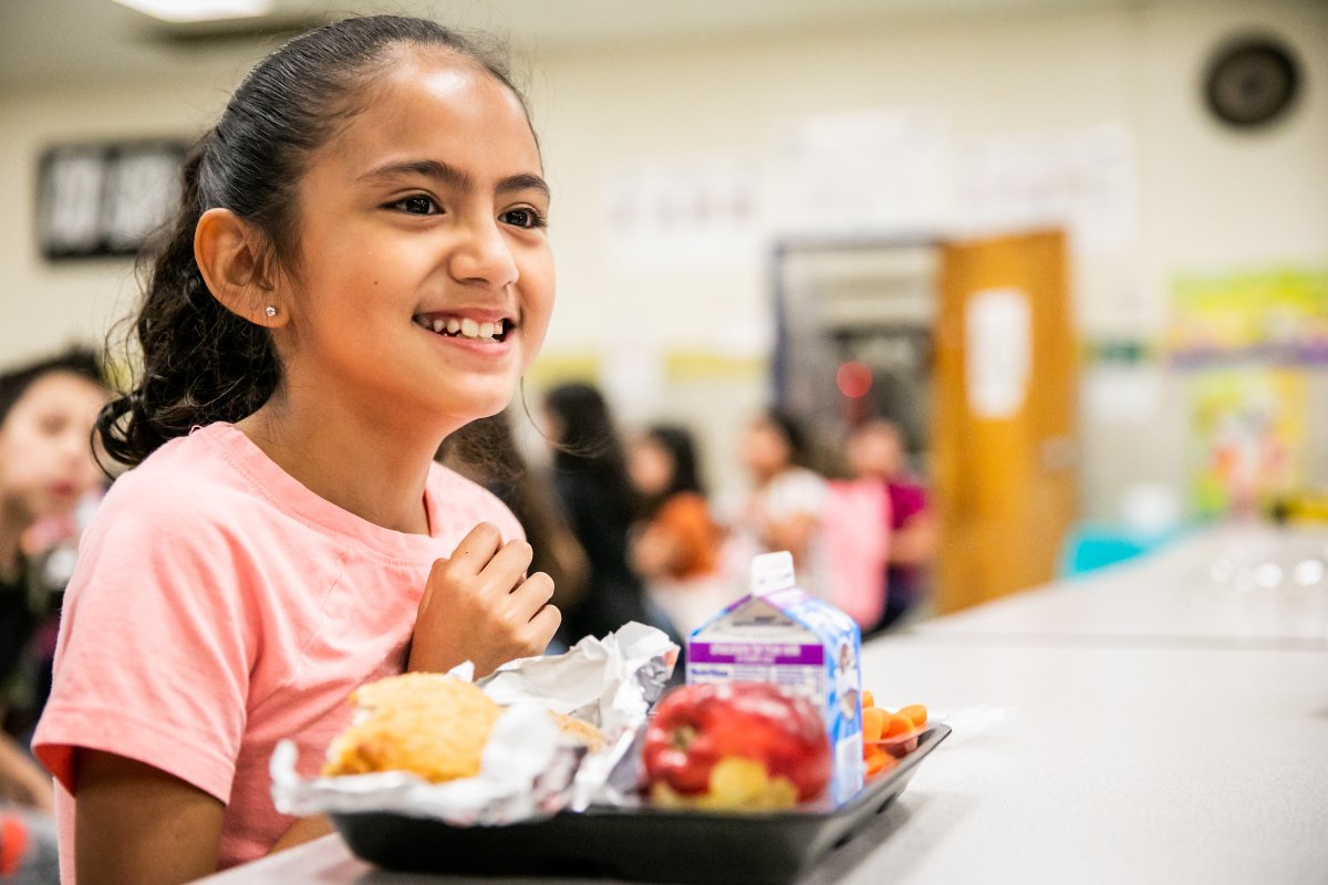 A girl with a ponytail sits at a school cafeteria tale with a lunch tray in front of her, including a carton of milk, a red apple, and other food. She is smiling and wearing a pink t-shirt
