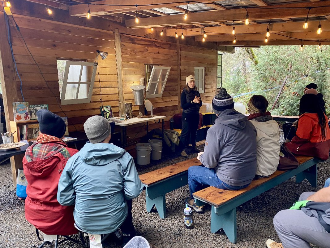 A group of people sit in a shedlike indoor/outdoor area in the woods wearing beanies and jackets listening to a woman explain how to tap bigleaf maple trees