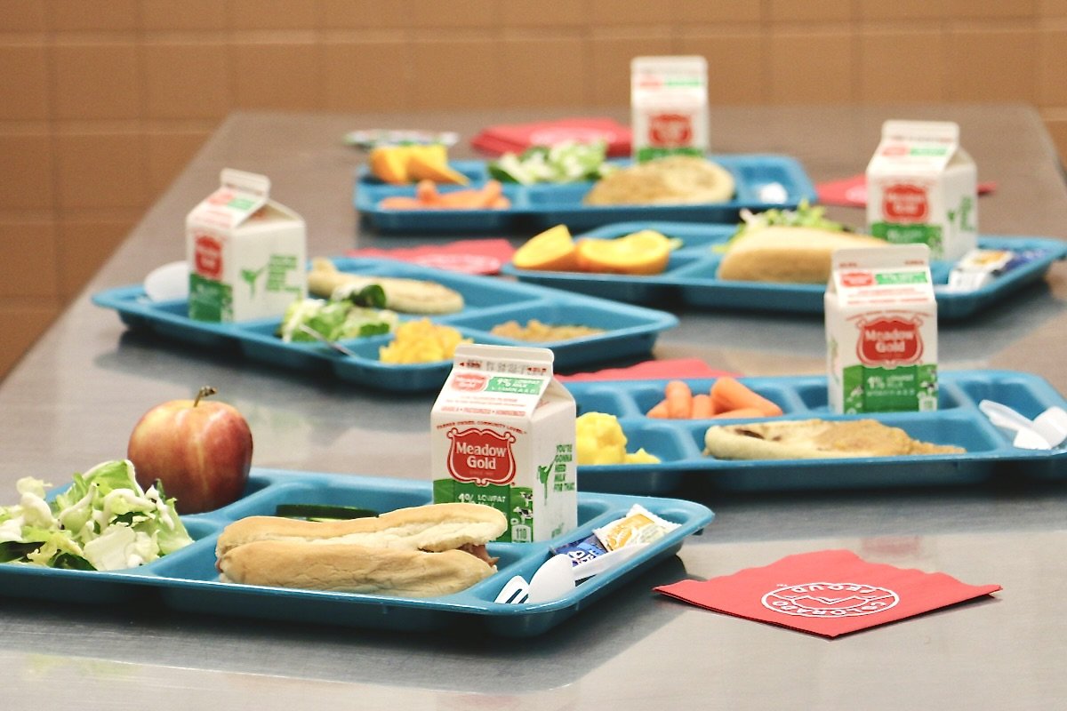 several trays of school lunches that incldues a salad, milk carton, and other items on a cafeteria table